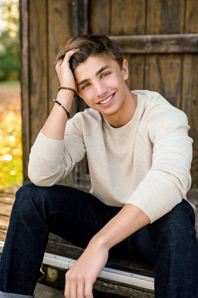 senior guy wearing light colored shirt with hand in hair leaning on barn while sitting