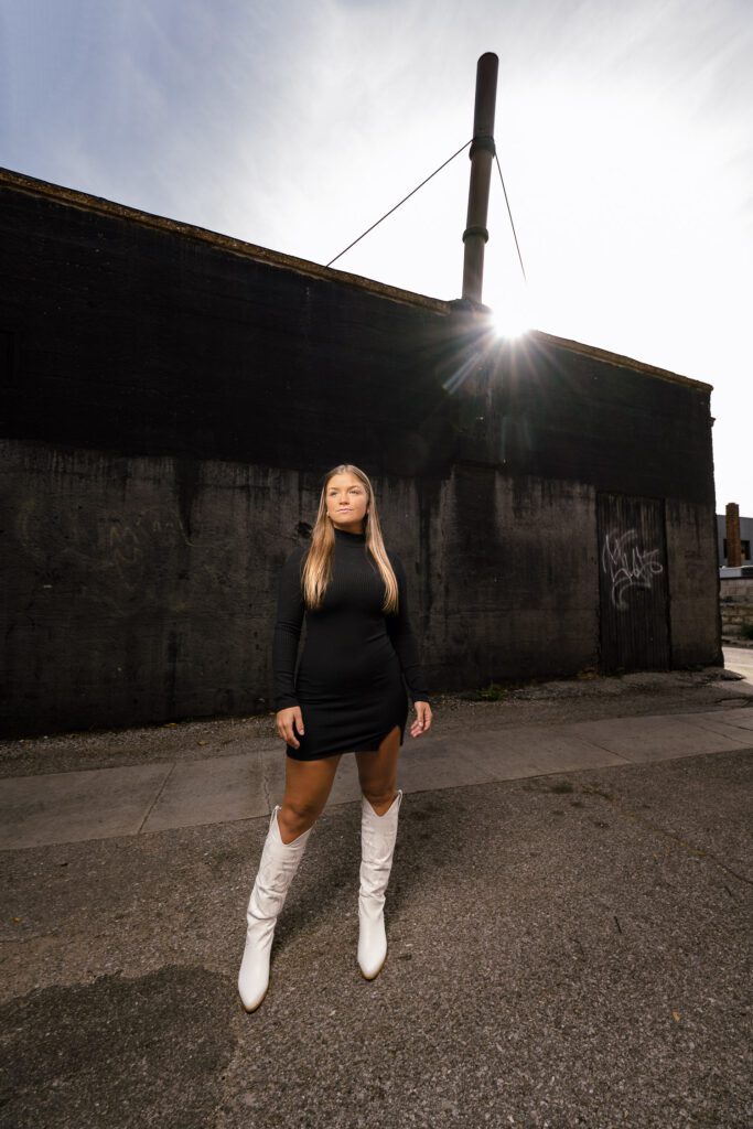senior girl wearing black dress and white boots stands in alley with sun shining