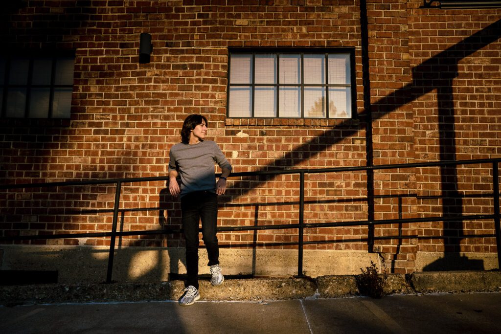 Columbia missouri high school senior guy leans on railing in front of brick wall with warm sunlight