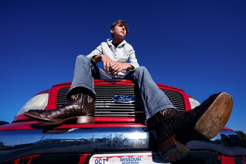 Ashland Missouri senior guy wearing blue jeans and boots sits on hood of red ford truck