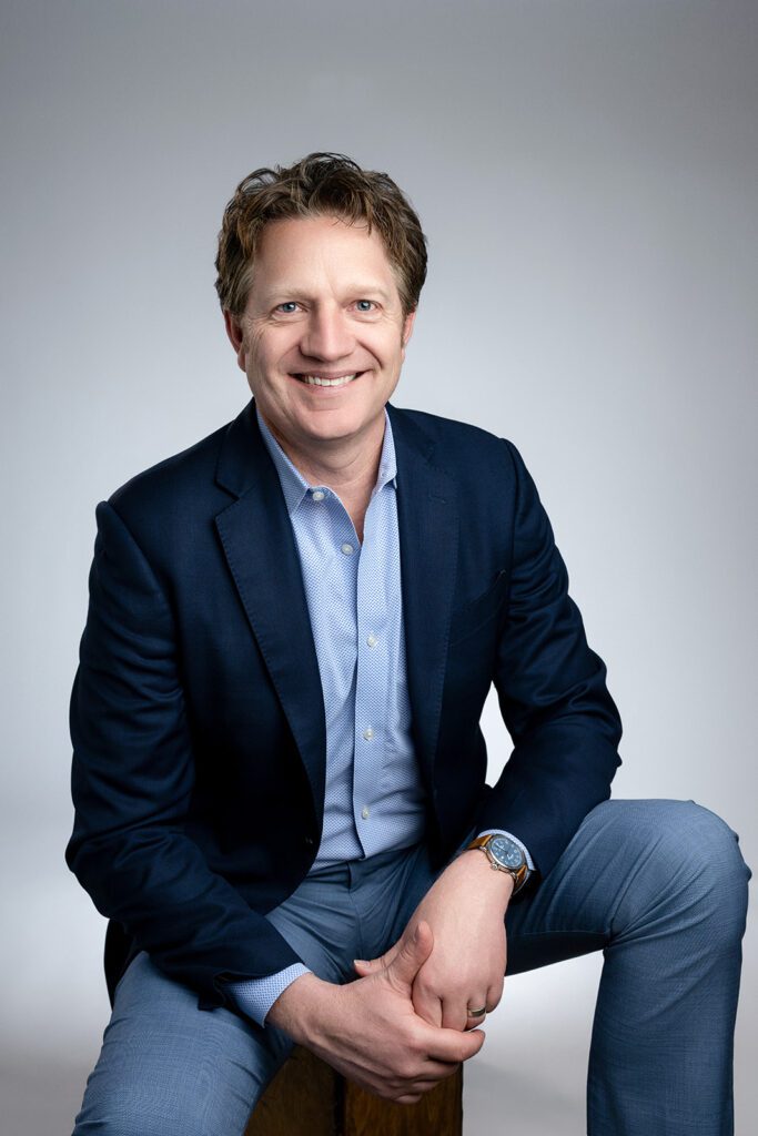 Male Business Executive wearing blue shirt and sport coat smiling with gray background