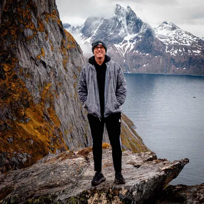 Guy standing by lake and white capped mountains wearing gray jacket.